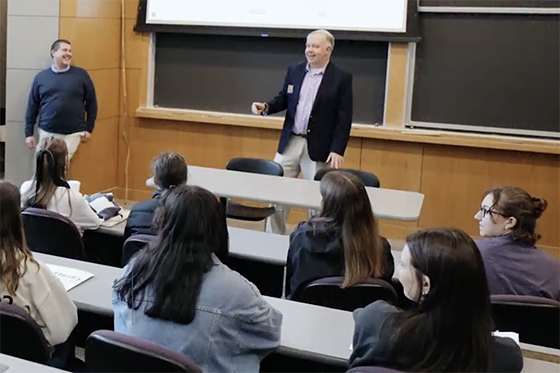 A professor speaks to a group of students seated in a lecture hall, while another professor st和s nearby smiling